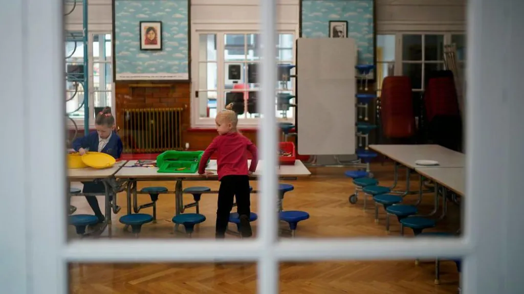 The photo is taken through a white window frame and shows two young pupils sitting at tables playing. One is wearing a red uniform and the other is wearing a blue uniform. 