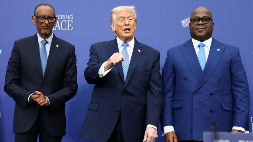 Paul Kagame, Donald Trump and Félix Tshisekedi stand next to each other. Kagame has his hands clasped in front of him, Trump is talking, looking to his left and holding his left fist in the air in front of his chest, and Tshisekedi appears to be standing to attention.
