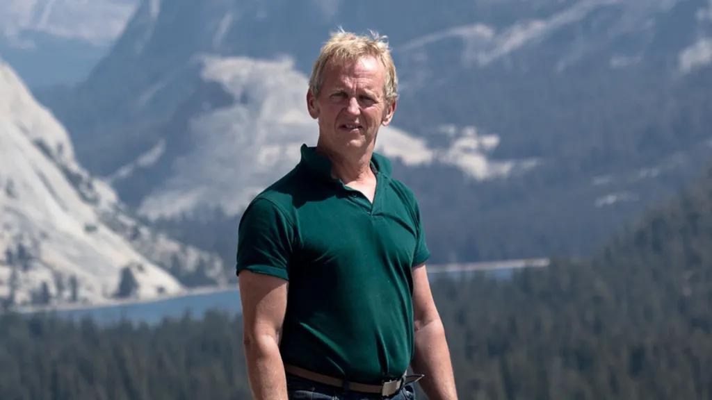 Stephen Lismore wearing a green polo shirt and jeans. He is standing in front of a backdrop of mountains and trees