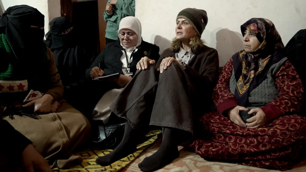 Hind Kabawat sits on the floor between two women, listening as she meets  residents of a refugee camp. She is wearing a dark coloured woollen hat, dark, wide-leg trousers and a light shirt and dark jumper. The women on either side of her are wearing headscarves, one is her assistant who is taking notes.