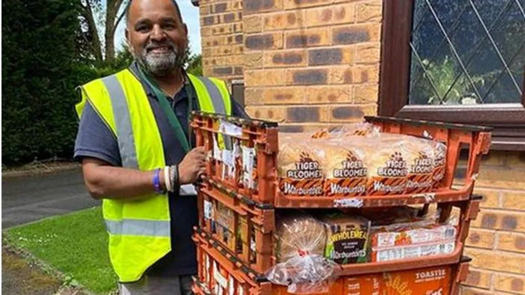 A man wearing a green high-vis jacket next to crates of bread. The crates are orange and behind him is grass and a brick wall of a house. 