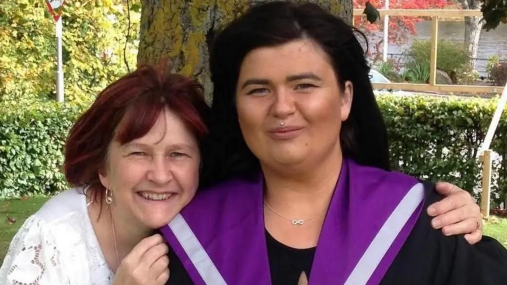 Jill and June after a graduation ceremony. Jill, on the left, is wearing a white lace top and June is wearing a black and purple graduation gown. They are standing in front of a tree.