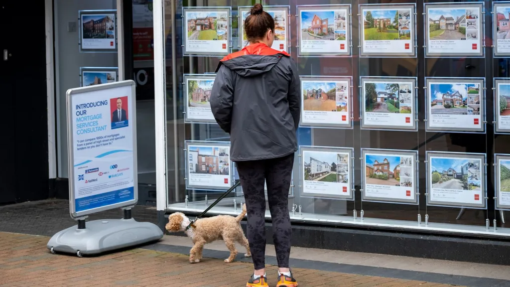 A woman looks in teh window of an estate agent . She is wearing sporty clothes and has a small dog on a lead with her