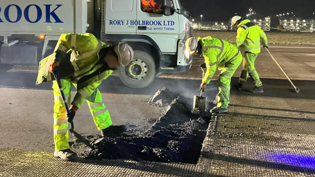 The picture shows a well-lit airport runway at night with two construction workers in yellow high-visibility clothing digging asphalt with shovels. A third worker walks behind them. In the background a large construction lorry can be seen and in the distance a multi-storey car park.