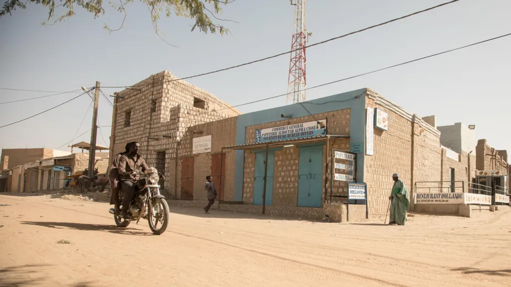 A man rides a motorbike along a dusty road in Timbuktu. Buildings made from concrete blocks line the street