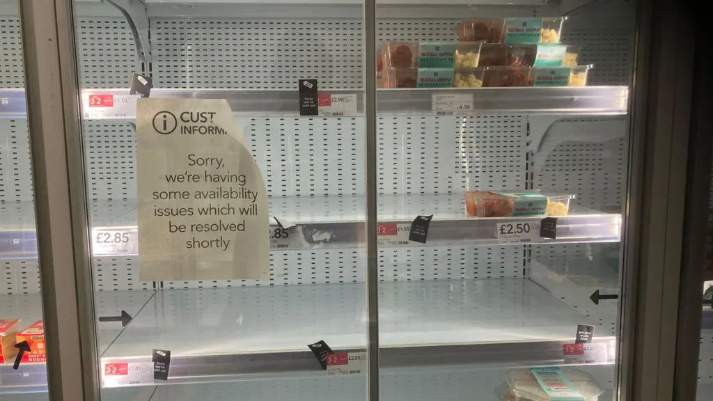 A shop fridge with almost empty shelves, and a sign stuck on the window which says "Sorry we are having some availability issues which will be resolved shortly"