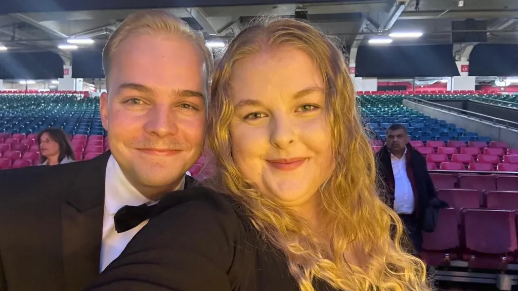 Ellis Pears wearing a tuxedo, with a white shirt and black bow tie. He is stood next to a woman with long wavy blonde hair. There are rows of seats behind them in a hall.