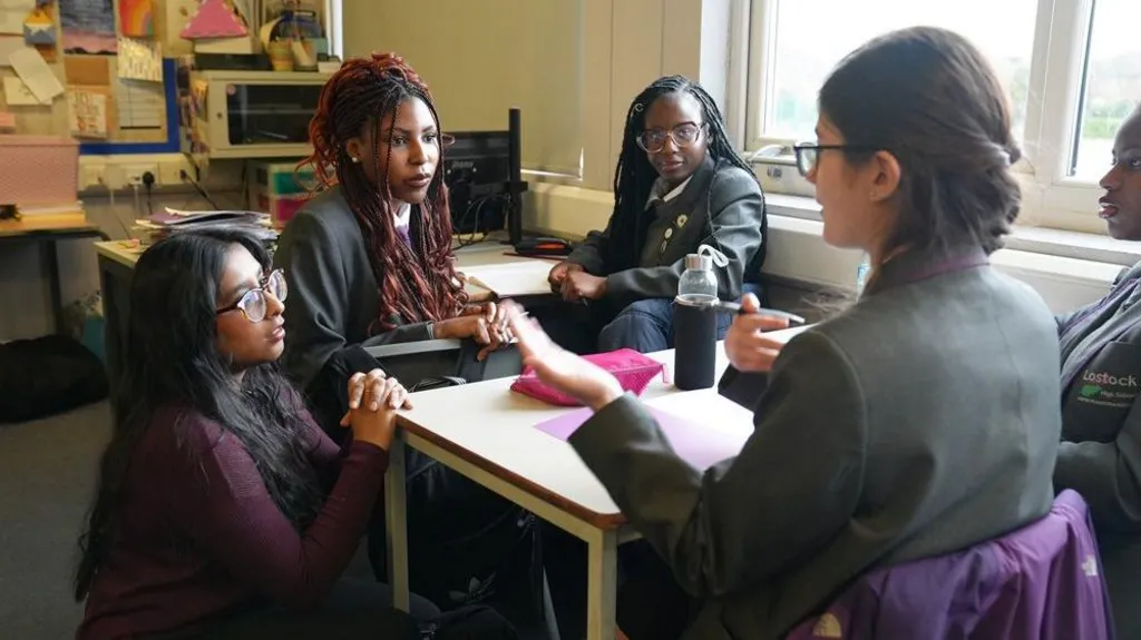 A teacher, with long back hair and glasses kneels down beside a school desk talking to some students . The students are wearing black blazers and white shirts. 