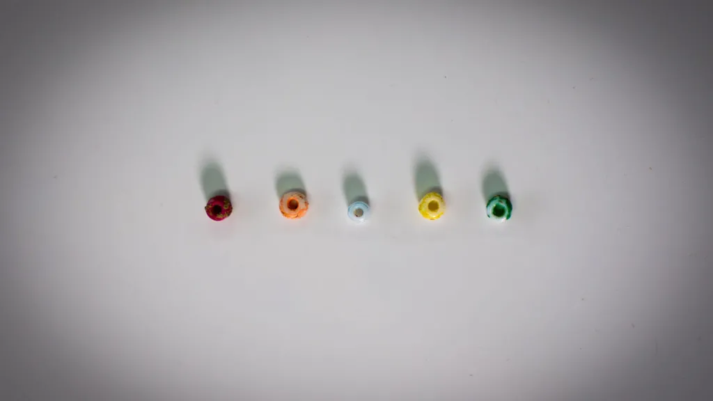 Five small round beads arranged horizontally on a white surface.  Each bead has a hole in the centre.  They are multicoloured, from left to right, they are dark red,  orange, light blue, yellow and green. 