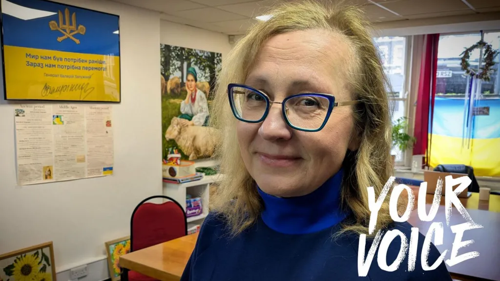 Larysa stands in a brightly-lit community room decorated with Ukrainian colours. Behind them, a large blue‑and‑yellow sign and an information board are mounted on the wall. A painting showing a woman with sheep is displayed nearby. Sunflower artwork rests on the floor against the wall, and red‑framed windows at the back are draped with a large Ukrainian flag. Tables and chairs are arranged around the room.