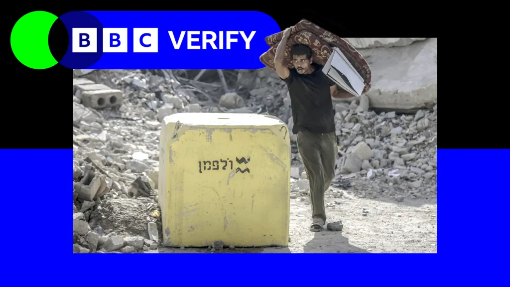 A man carrying a folded mattress in Gaza walks past a yellow block placed by the Israeli military, with rubble visible in the background. The image has been imposed over the BBC Verify logo and colours. 