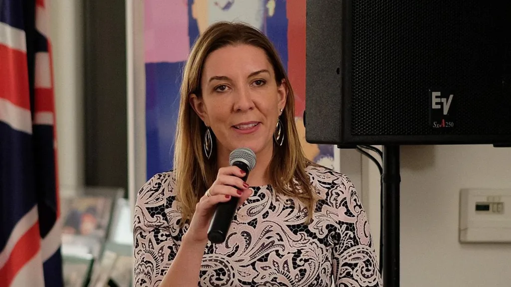 Dame Antonia Romeo speaks into a microphone in front of a Union Jack flag.