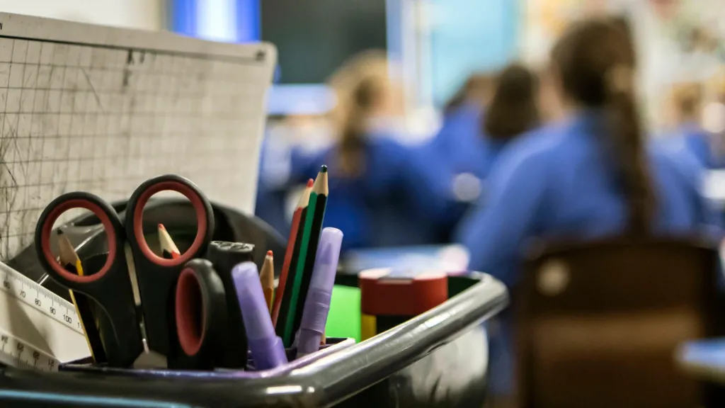 A stock image of burry schoolchildren in the background with a pot full of pens and other stationary in the foreground