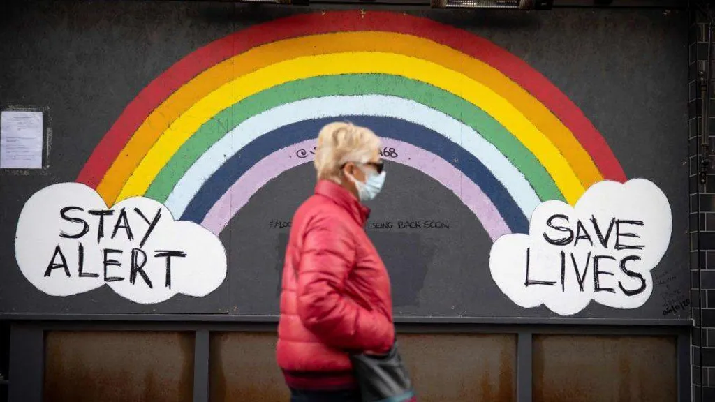 A woman wearing a face mask, sunglasses and a red coat walks past a rainbow mural on a wall which reads "Stay alert save lives" during the Covid pandemic.