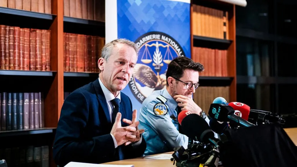 Etienne Manteaux speaks to the press with many microphones infront of him and a man in a police uniform sitting next to him. There is a big bookshelf behind them with many books.