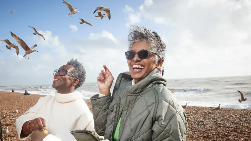 Two women smiling whilst eating fish and chips on a British pebble beach. Seagulls surround them and the sea is in the background.