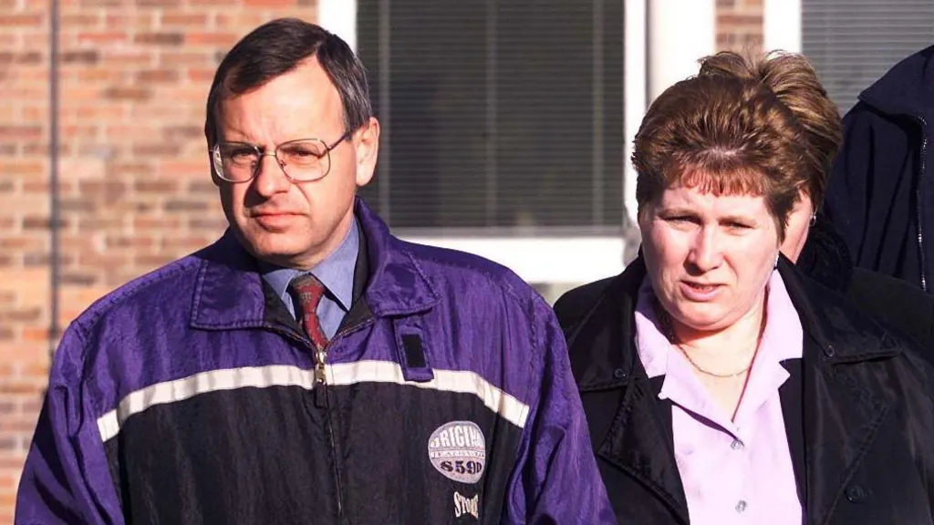 Graham Hall is wearing a shirt and tie with a purple, blue and white anorak over it. He has parted hair and square-shaped glasses. He is walking alongside Lorinda Hall, who wears a pink blouse and neck chain under a black jacket. She has backcombed hair.  Behind them is a brick court building.