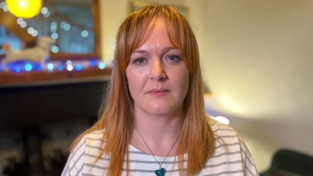 A woman with ginger hair and a nose ring looking at the camera. She is not smiling. She appears to be standing in a living room but the background is blurred. She is wearing a white and grey stripy top and a blue heart necklace.