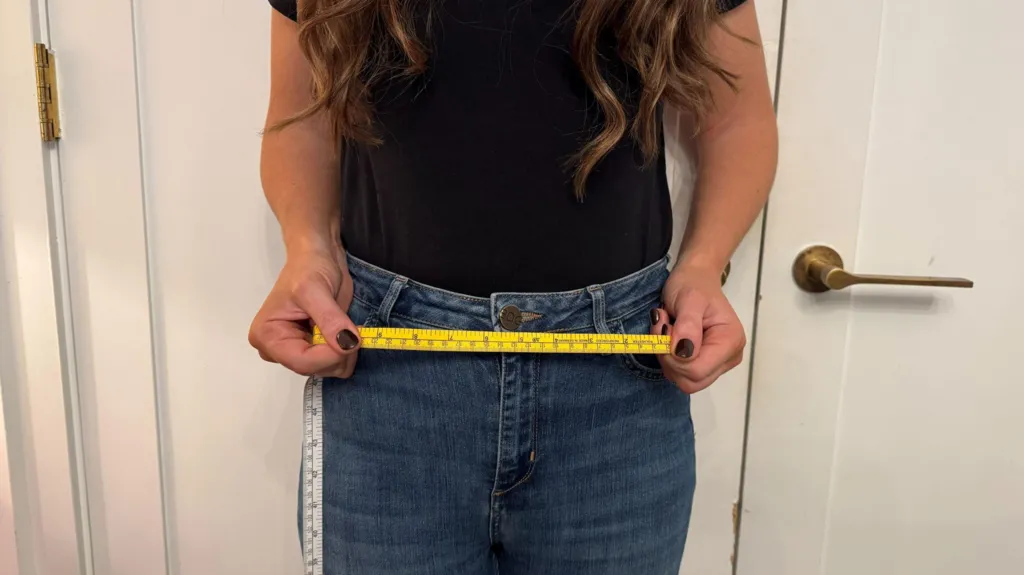 A woman in jeans stands with a yellow tape measure across her waist.