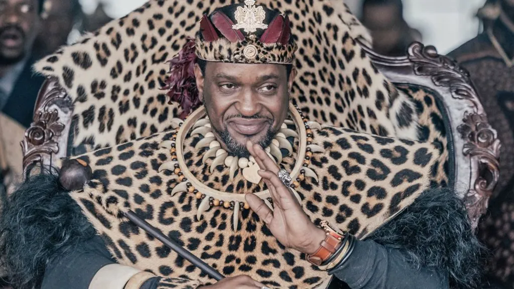 Misuzulu kaZwelithini sits on a throne and waves in his royal regalia, including animal skins and black ostrich feathers. His jewellery includes a silver lion ring and necklaces that include animal teeth.