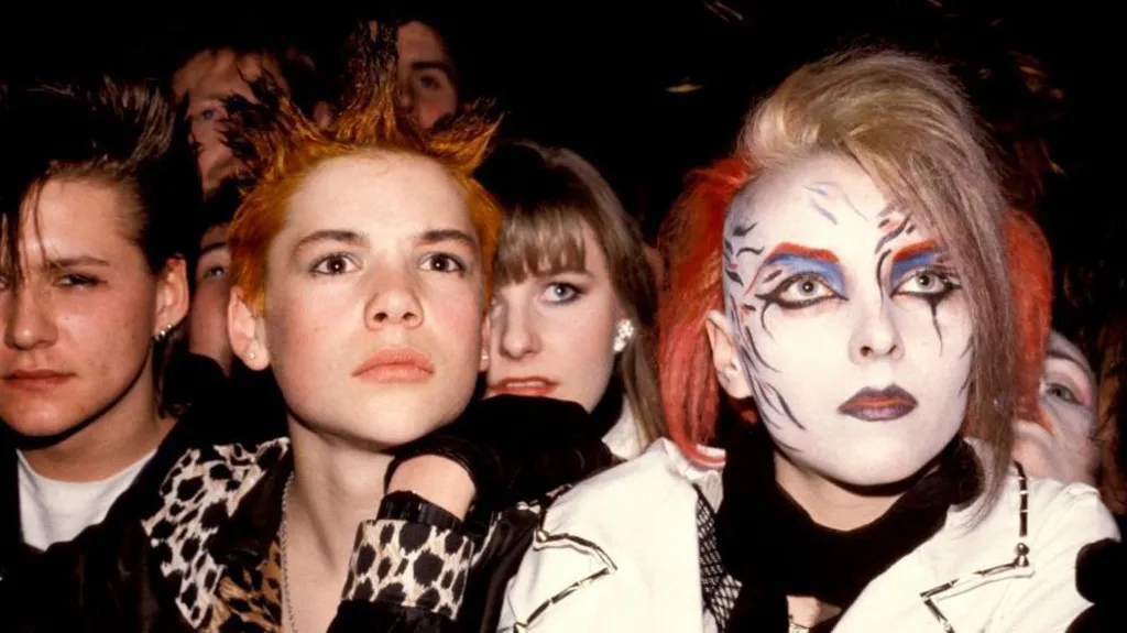 Two young women from the early 1980s photographed facing the camera. One has blonde and red hair with elaborate make-up of a white face, red eyebrows, blue eye shadow and blue-grey lips  