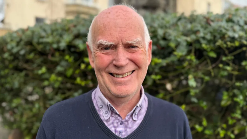 Councillor Michael Harriott smiles at the camera. He has white thinning hair. He wears an open-necked checked shirt and a navy v-neck jumper.  He is standing in front of a hedge.