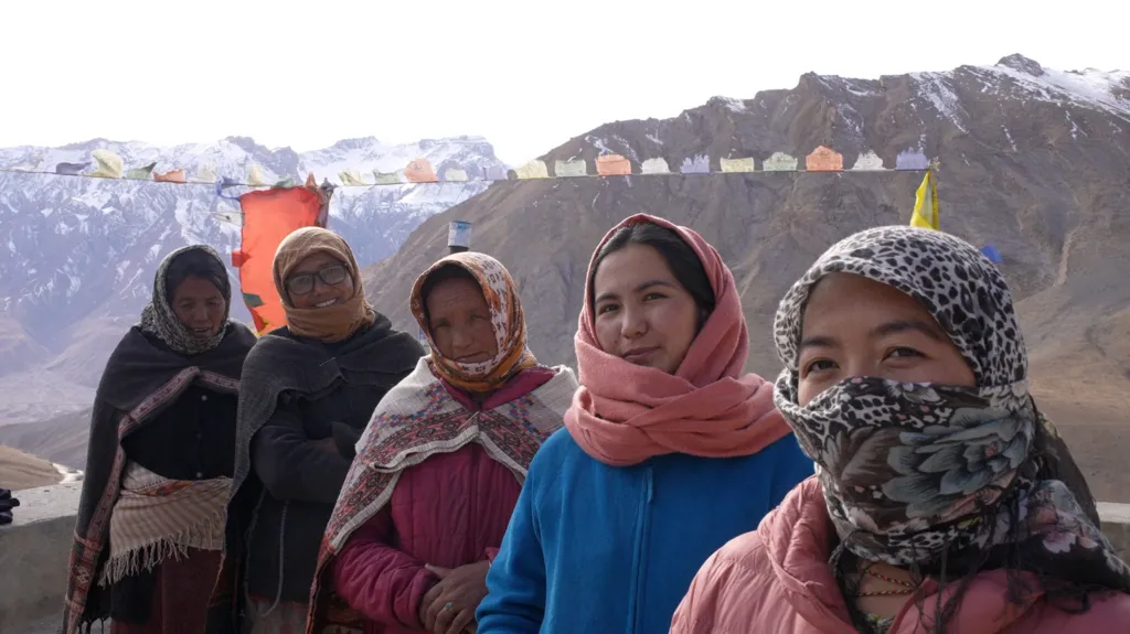 Five women wearing winter jackets look into the camera. They are standing in front of snow-covered peaks.