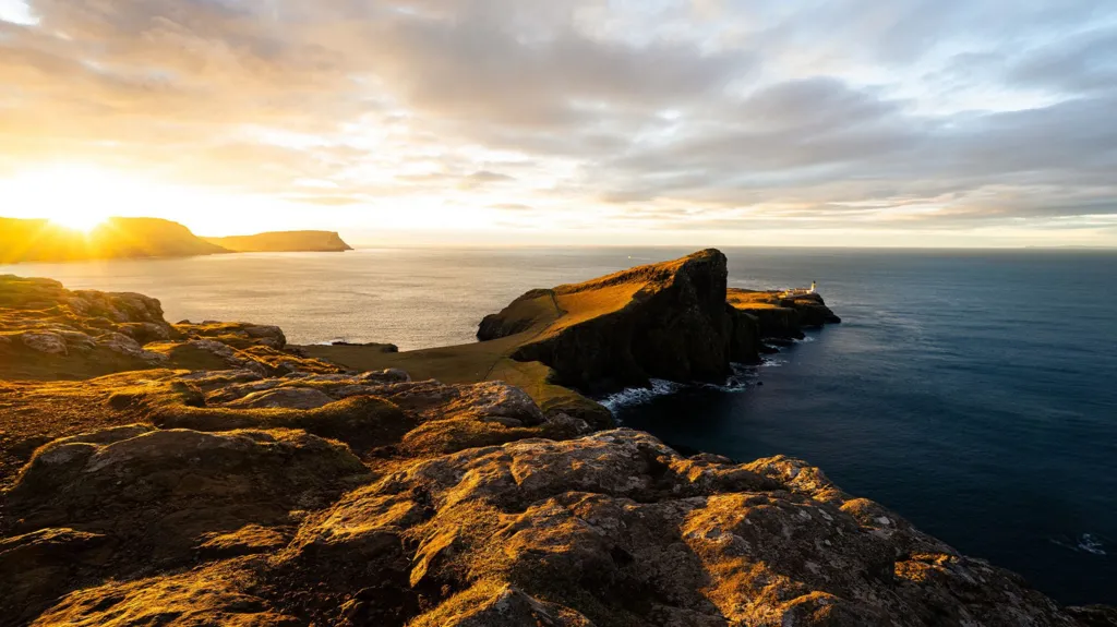 A dramatic coastal headland at sunset, with golden light hitting rocky cliffs that extend into the sea, and a lighthouse visible at the far end of the peninsula.