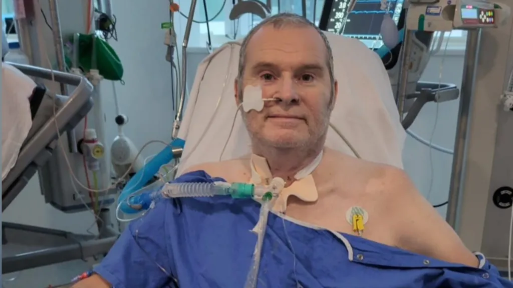 A man in a blue hospital gown sits in a chair and looks at the camera. He has a breathing tube in his nose and other tubes taped to his throat. Medical machines can be seen behind him.