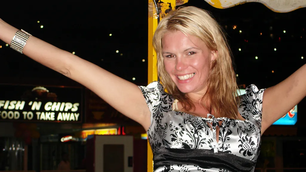 Kirsty Wilkinson wears a silver short sleeved dress with a pattern of black flowers on it. She is white, with bleached blonde hair which comes below her shoulders. She has her arms raised and is smiling at the camera. She wears a wide silver bangle. In the background, which is at night, a sign reading "Fish'n'Chips food to take away" is illuminated. There is a yellow pole behind her and the edge of a canopy. It looks like a fairground site.