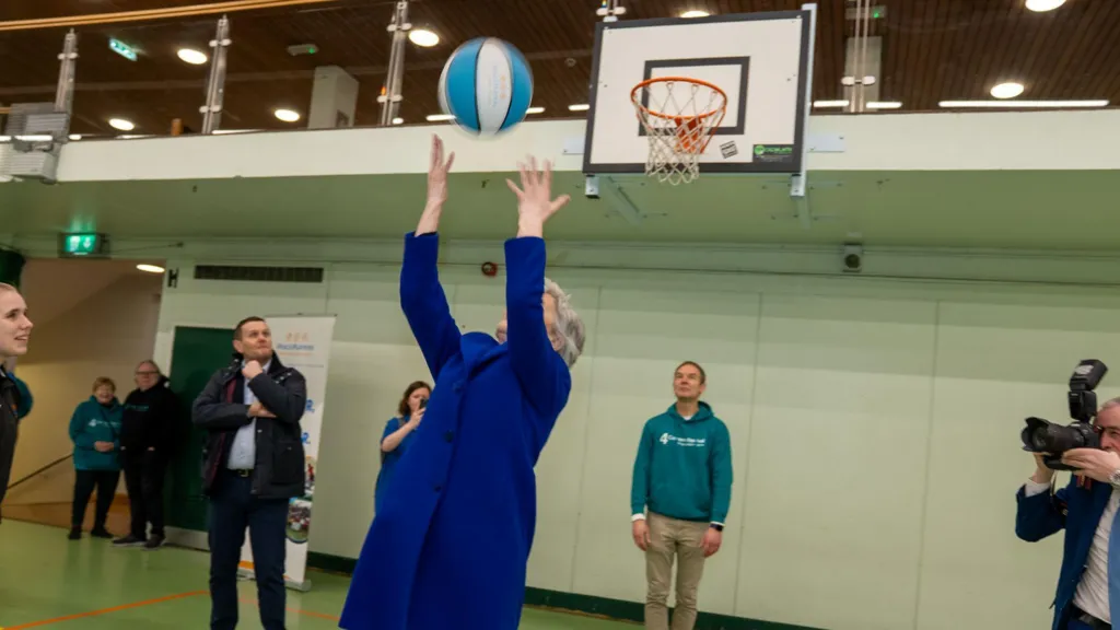 Catherine Connolly in a blue jacket throwing a blue and white basketball over her head towards a basketball hoop. 