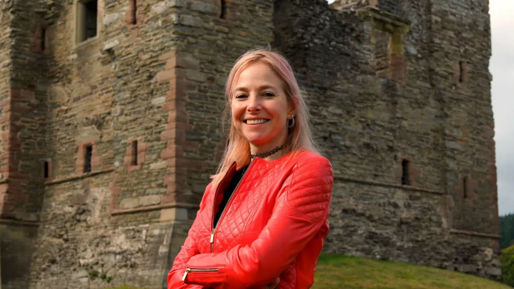 Prof Alice Roberts standing in front of the ruined castle keep. She has pink tinted hair and is wearing a red jacket. Her arms are folded and she is smiling broadly. 