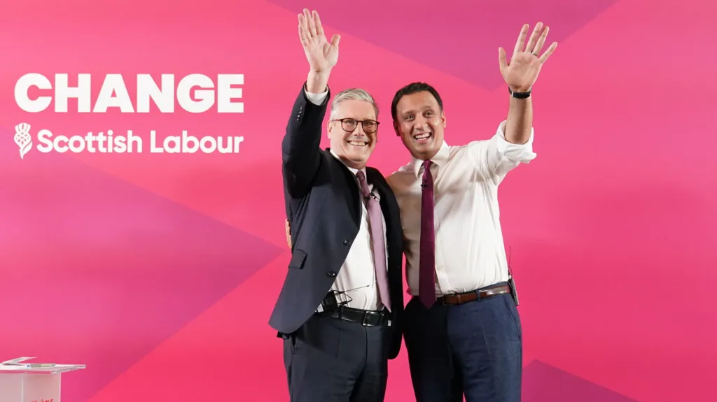 Sir Keir Starmer, left, wearing a dark suit over a white shirt and a pink tie. Anas Sarwar, right, wearing a white shirt with a red tie and dark blue trousers. Both are waving while standing in front of a red background.