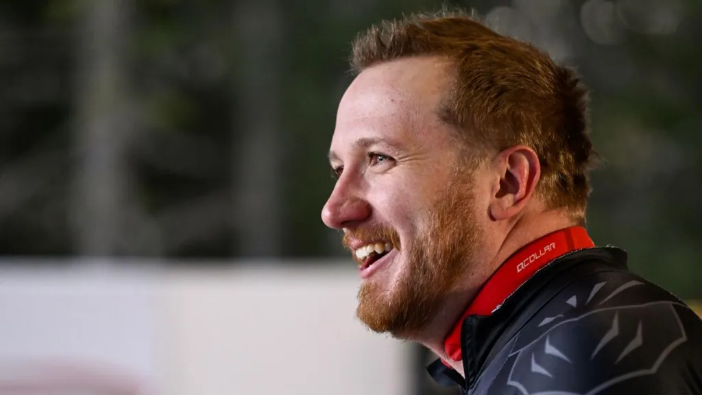 A man wearing a black, form‑fitting athletic suit with geometric panels stands outdoors in a snowy setting. The man faces sideways, looking toward someone off‑camera, with blurred winter trees and a partial event banner in the background.