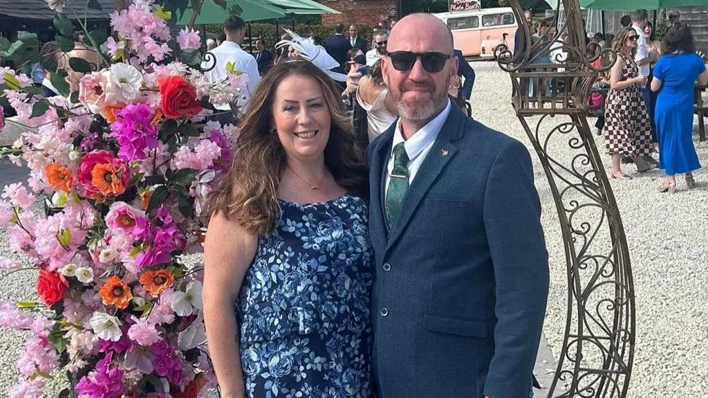 Jemma Williamson, a woman with wavy brown hair in a blue flowery dress is smiling at the camera and standing next to a man in a blue suit with a green tie and sunglasses. Behind them in a flowery display 