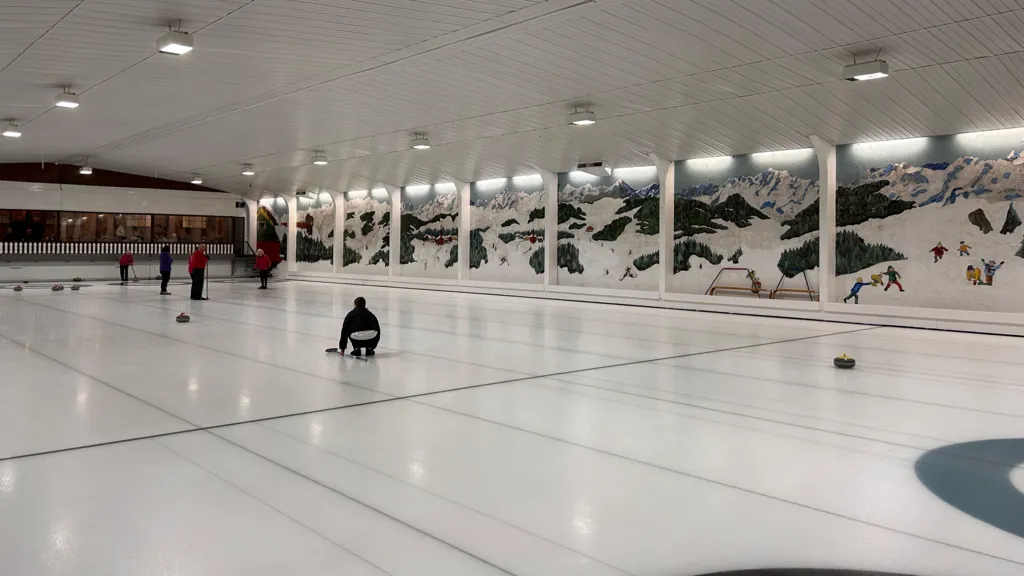 A white ice-rink with some curlers playing, showing a mural of an alpine scene along the length of the back wall.