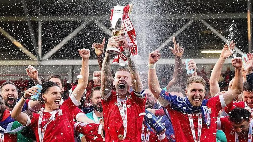 Wrexham AFC on the pitch for their promotion celebration. Player holding a trophy, with champagne thrown 