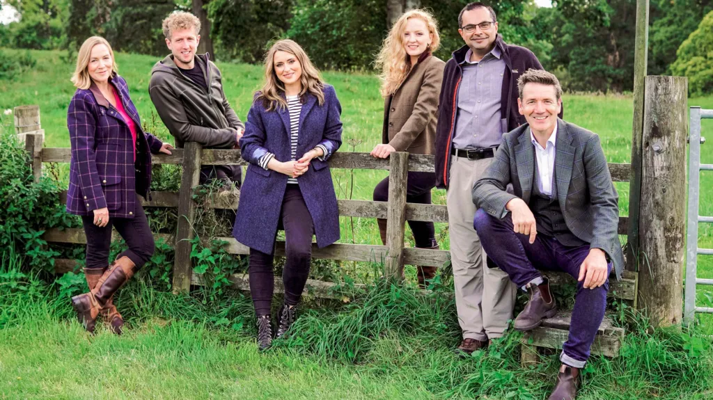 Landward presenters Arlene Stuart, Cammy Wilson, Anne McAlpine, Rosie Morton, Shahbaz Majeed, Dougie Vipond, dressed in fashionable country clothes, pose around a fence in a lush green field