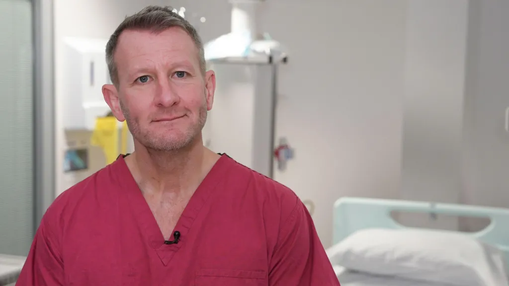 A white man with brown hair in red hospital scrubs sits in a hospital room in front of a hospital bed 