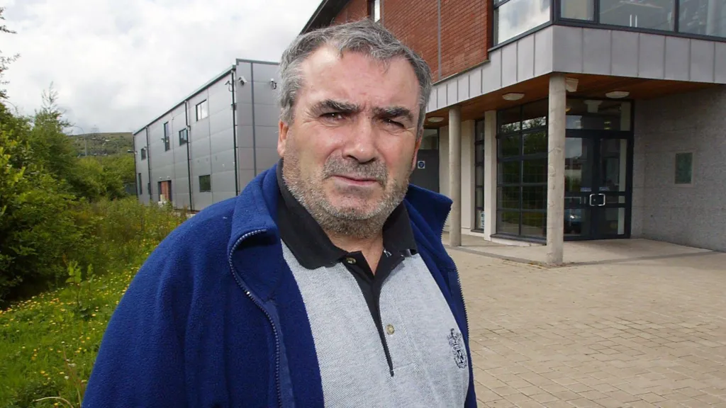 Freddie Scappaticci pictured in west Belfast in 2003 before he went into hiding in England. He is wearing a grey polo shirt under a blue fleece jacket.
He is looking into camera and has greying short hair and grey stubble.  He is standing in front of a red brick building with panels of grey cladding.