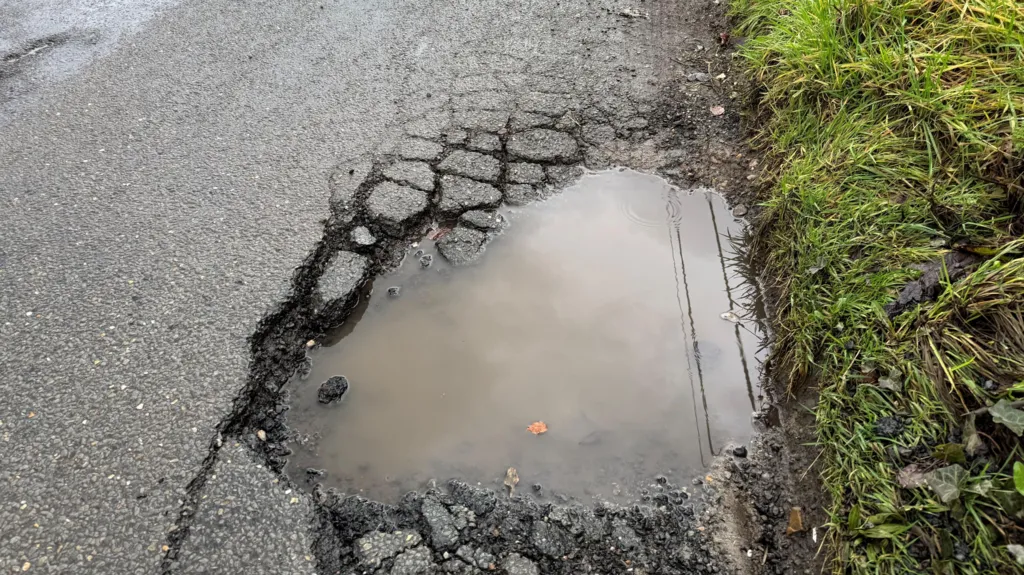 A large water-filled pothole on a road and next to a grass verge. There are cracked bits of tarmac all around it.