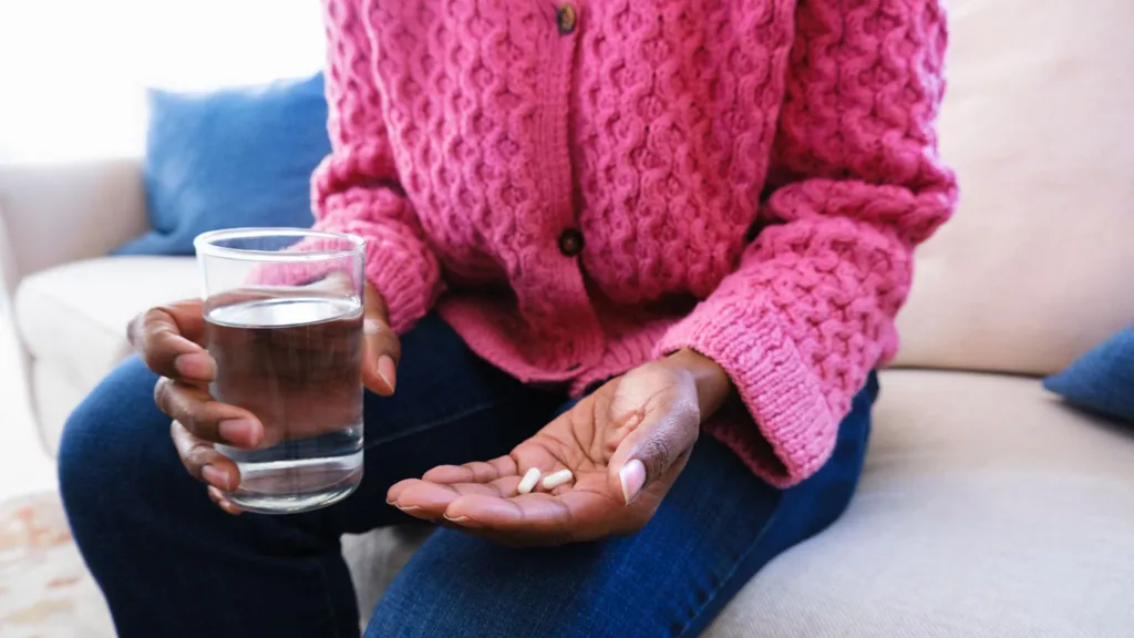Woman wearing bright pink cardigan and dark blue jeans hold two pills in one hand and a glass of water in the other