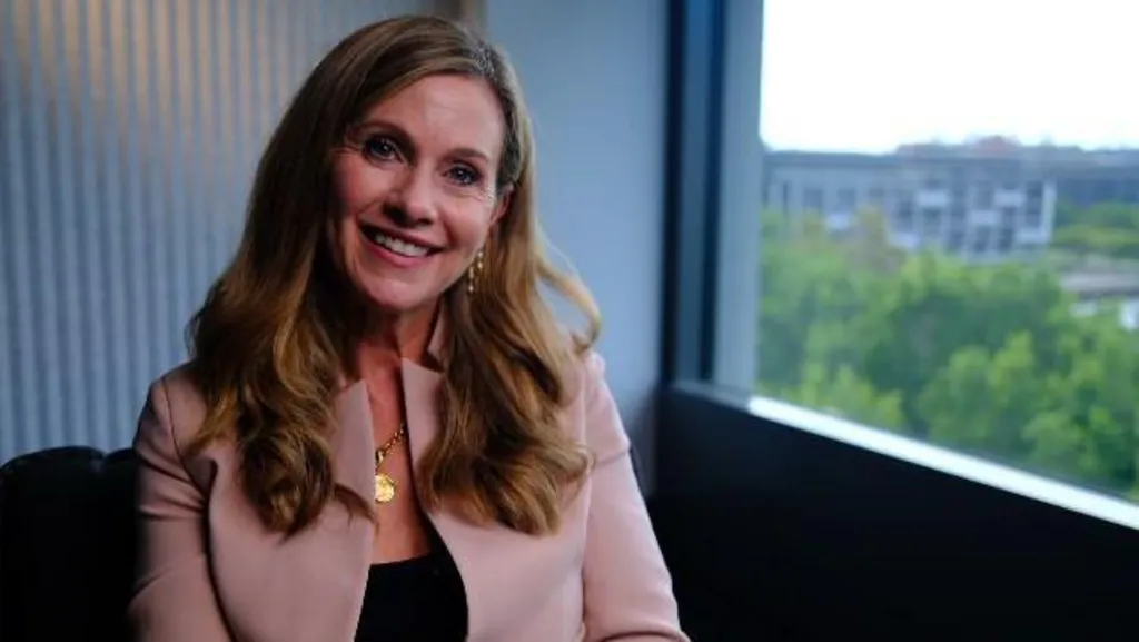 Julie Inman Grant in a pale pink jacket and black blouse smiles as she looks at the camera.  She is sitting next to a window. 