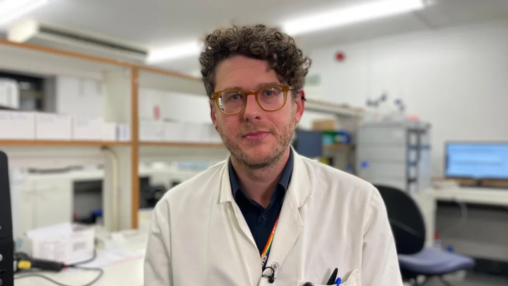 Dr Alex Lawson in his lab coat with an office in the toxicology lab in the background