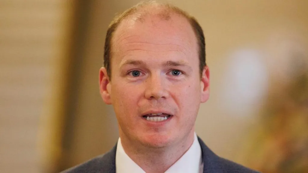 Close-up of a man inside a building, we can see the tops of his shoulders - he is wearing a dark suit and white shirt. He has balding short dark hair