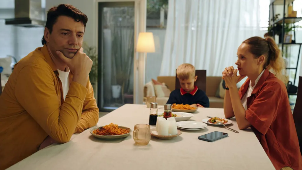 A man with a yellow shirt and a brown hair, a woman with blonde hair and a red shirt and a young child with blonde hair and navy jumper sat at a dinner table. The man and woman look aloof and the child is staring down at his plate. 