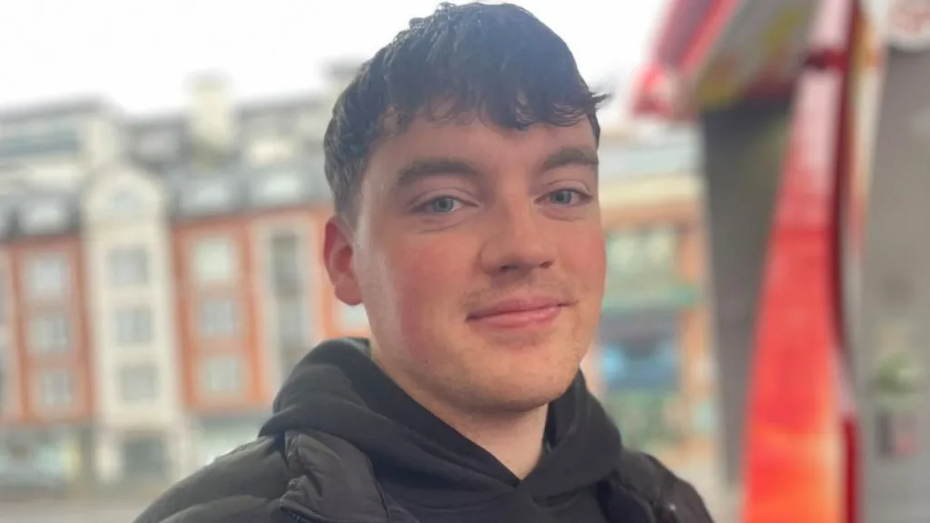 A man looks and smiles into the camera. He has mid length brown hair which has been pulled down over his forehead. He is wearing a black hoody and dark coat.