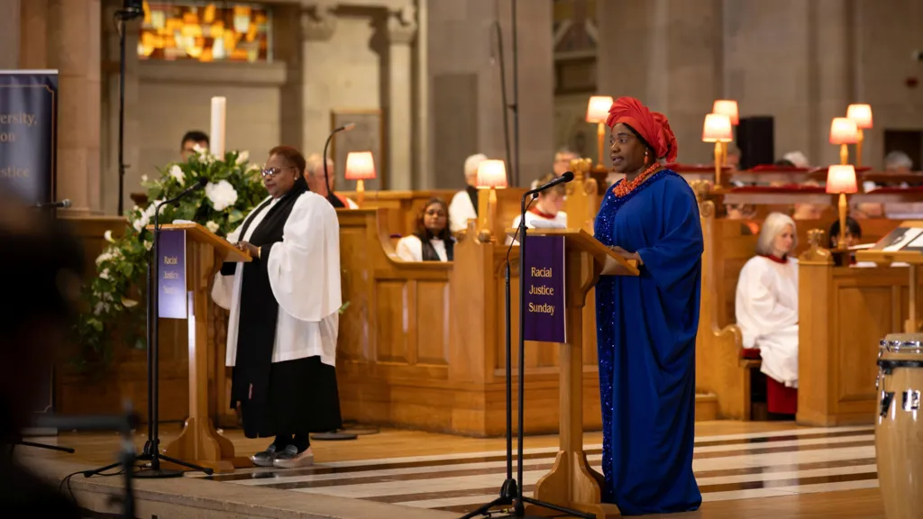 Two women are standing at podiums in the church. On the right a woman is wearing native dress and a red hair wrap. The dress is bright blue in colour. On the left, a woman is wearing a white robe and black scarf. 