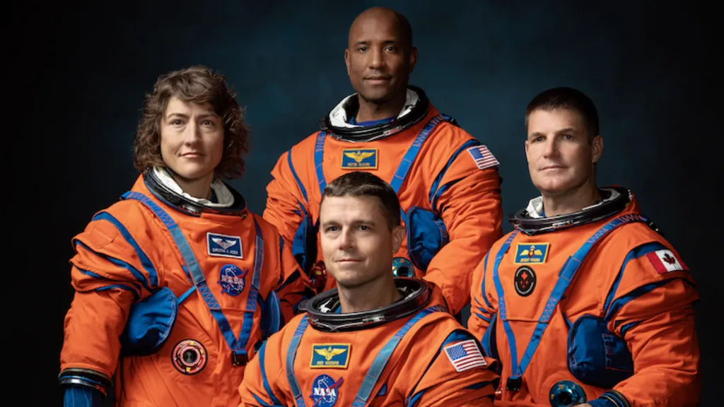 The image shows four astronauts in bright orange space suits posing for a formal group portrait against a dark background. The suits are bulky with blue joints, straps and pockets, emphasising their technical design. Three astronauts stand behind one who is seated, all facing the camera. They all have fairly neutral expressions, keeping the focus on the uniforms. Mission-style patches and flags, including US and Canadian flags, are visible on their arms and chests. Soft, focused lighting makes the vivid orange suits stand out dramatically.