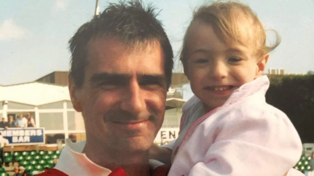 A man smiling as he holds his daughter in a photo. He has black hair and she has blonde hair and is wearing a pink top.